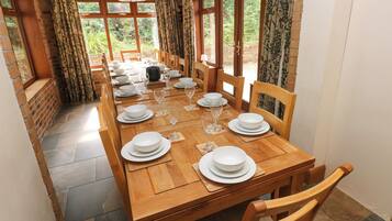 Uma sala de jantar com mesa de madeira posta para refeição, cercada por cadeiras, com vista para áreas verdes através de grandes janelas.