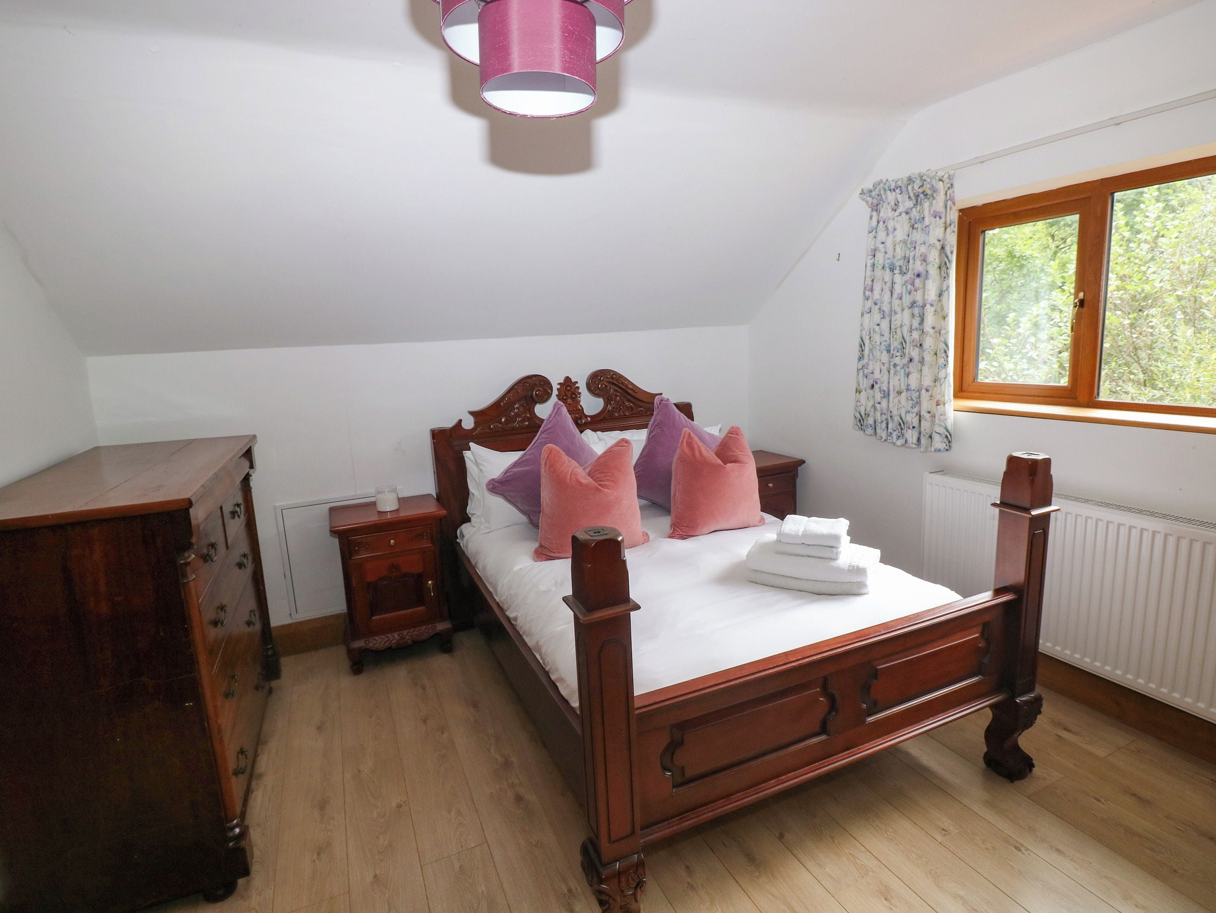 A bedroom with a wooden bed, a nightstand, a dresser, a window with curtains, and a radiator.
