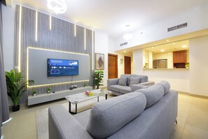 Ein modernes Wohnzimmer mit Flachbildfernseher, grauen Sofas, einem Couchtisch und einer Küche im Hintergrund.