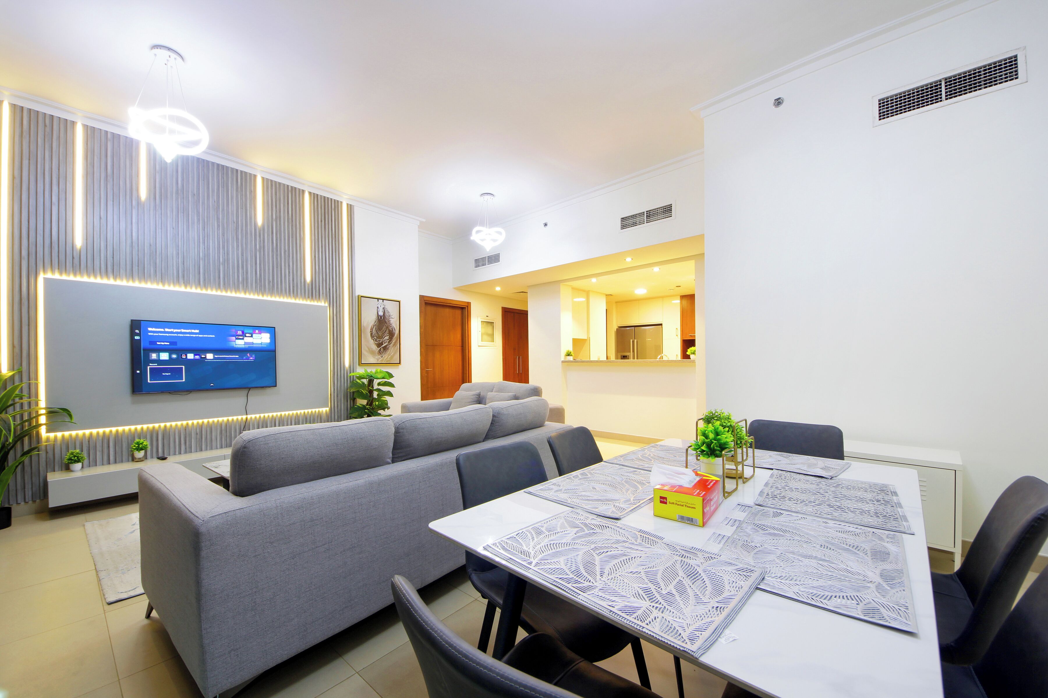A modern living room with a grey sofa, a dining table with patterned tablecloths, and a flat-screen TV mounted on the wall.