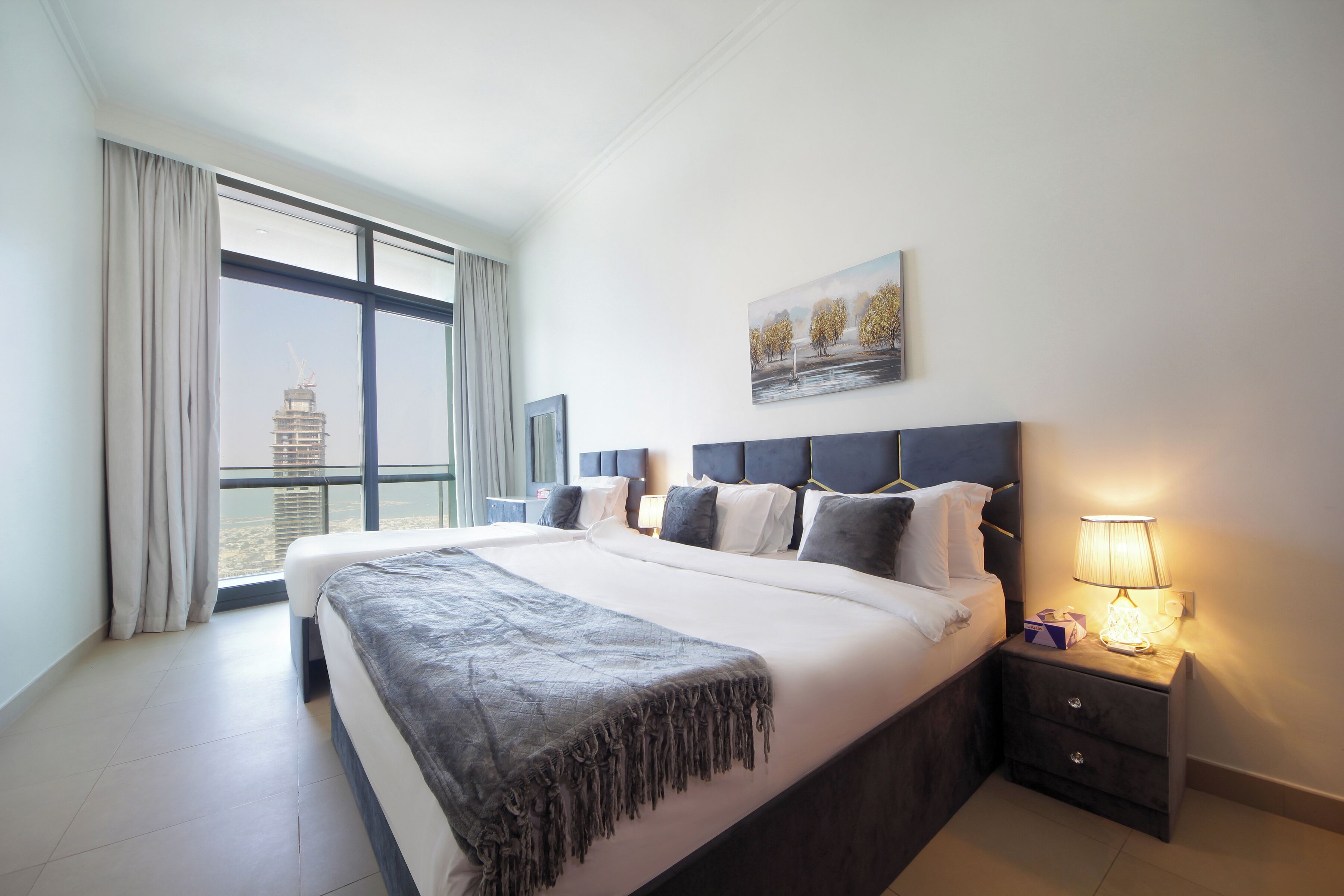 Quarto de hotel moderno com uma cama grande, uma mesa de cabeceira com abajur, vista para a cidade através de grandes janelas e um quadro na parede.