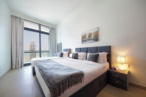 Ein modernes Hotelzimmer mit einem großen Bett, einem Nachttisch mit Lampe, Blick auf eine Stadtlandschaft durch große Fenster und einem Gemälde an der Wand.