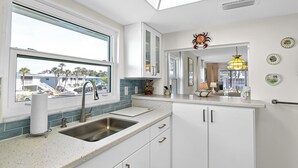 A modern kitchen with a large window, white cabinetry, and a stainless steel sink. - West Bay Cove 213 · 3 Blks to Beach · 30-Night (Holmes Beach)