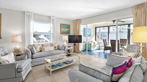 A modern living room with a sofa set, a coffee table with books, a flat-screen TV, and a balcony with outdoor furniture. - West Bay Cove 213 · 3 Blks to Beach · 30-Night (Holmes Beach)