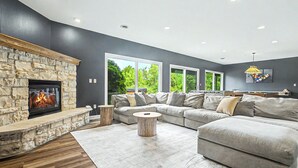 A modern living room with a stone fireplace, a sectional sofa, and large windows offering a view of greenery. - Grand Galena Hideaway [ Luxe Retreat w/ Hot Tub & Games ] (Galena)