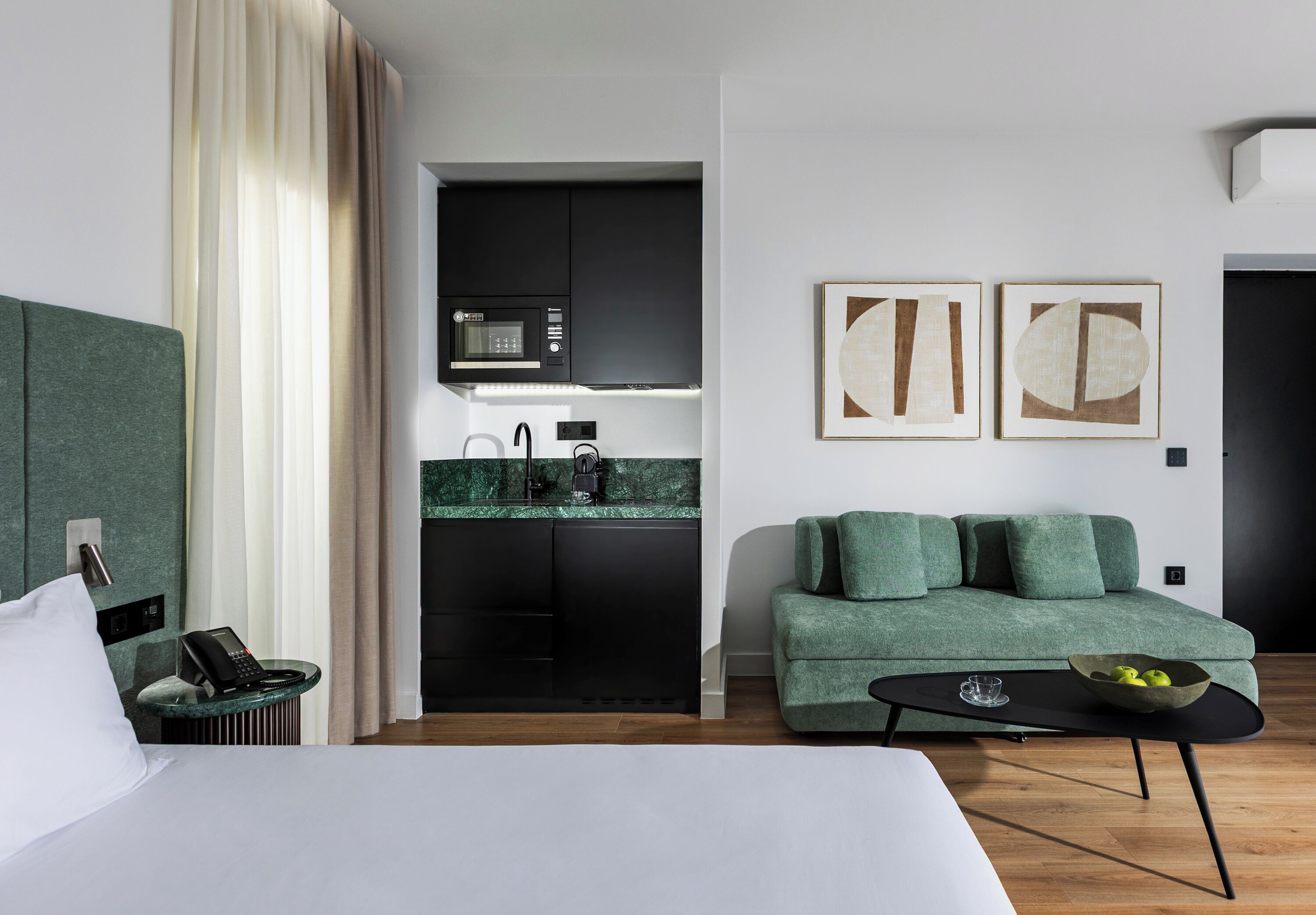 A modern hotel room with a bed, a green sofa, a small table, a kitchen area, and abstract wall art.