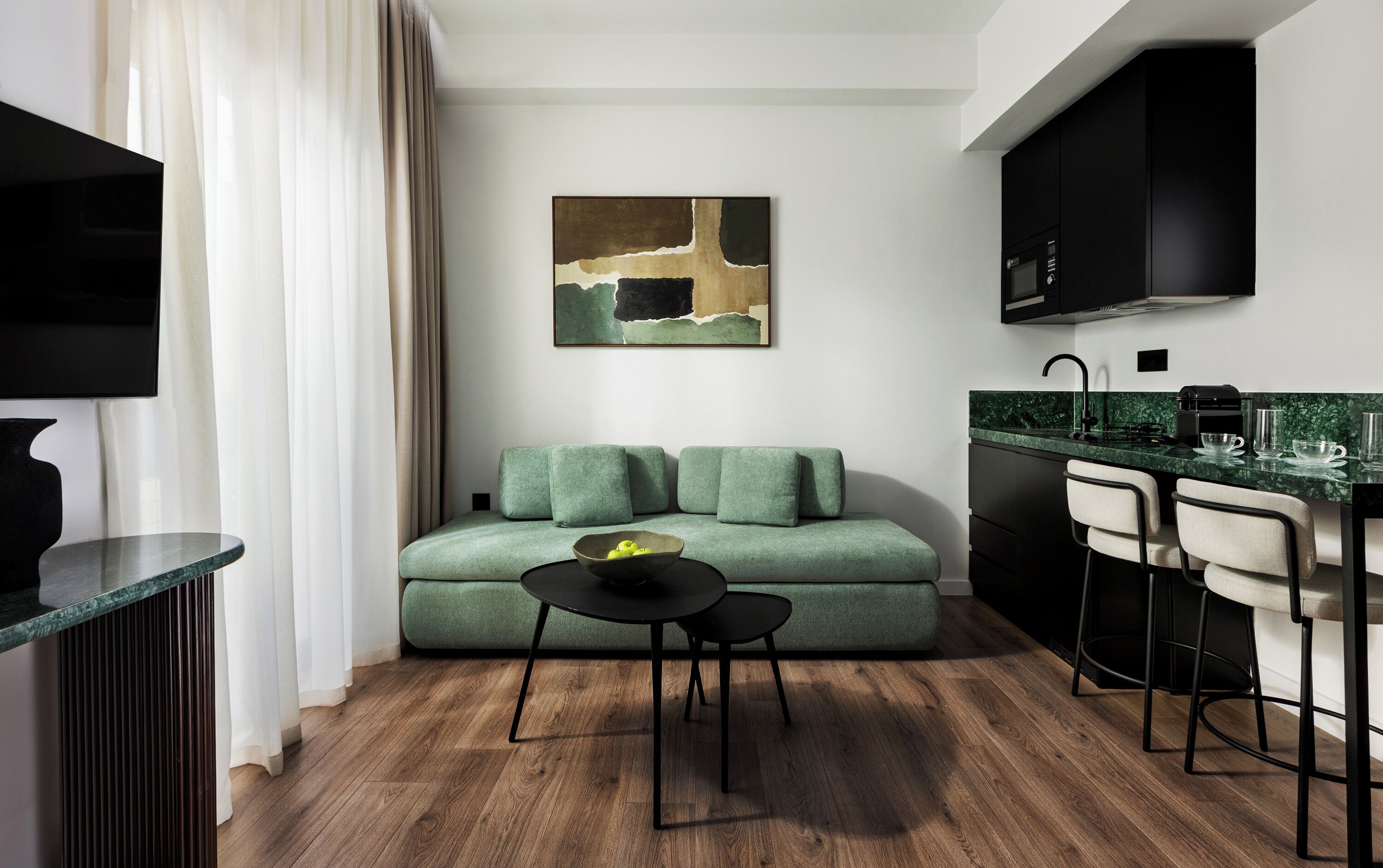 A modern living room with a green sofa, a small round table, and a kitchen area with a black countertop and bar stools.