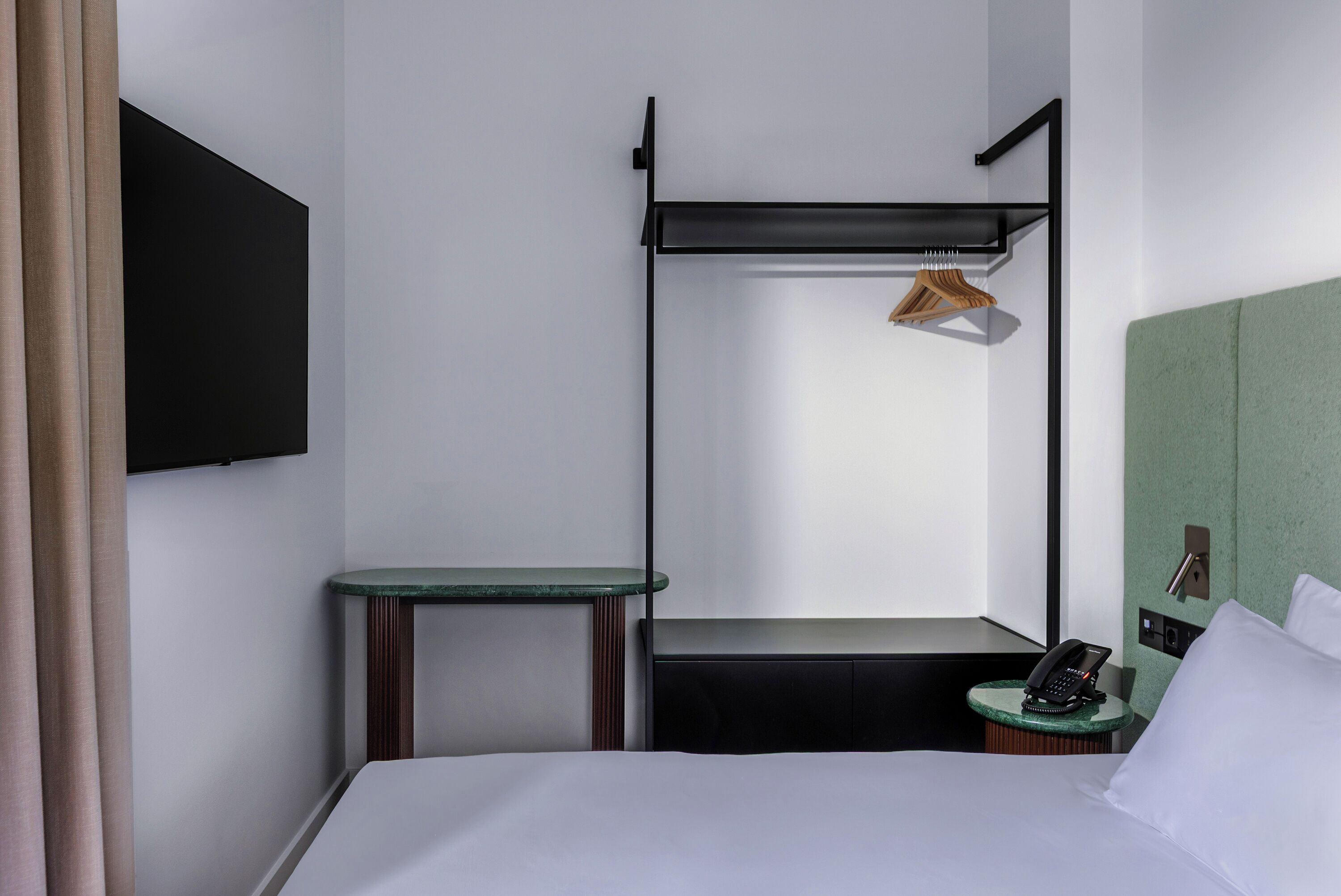 Una habitación de hotel con cama, una mesita, una banca, un televisor montado en la pared y un teléfono sobre una mesita auxiliar, además de una pared verde decorativa.
