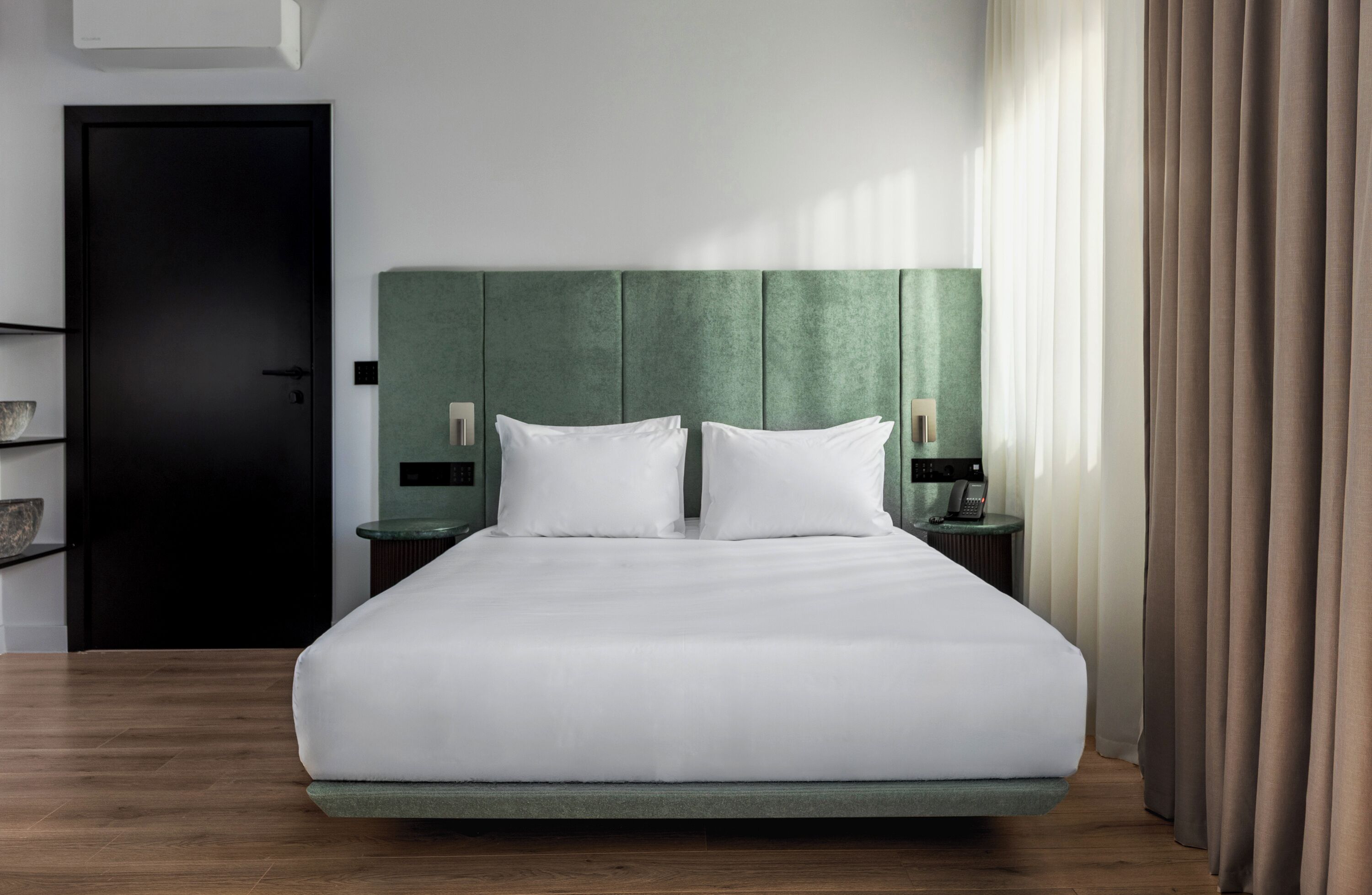 Ein Hotelzimmer mit einem Bett, einem Nachttisch, einem Schranken, einem grünen Kopfteil, einer schwarzen Tür und einem Fenster mit Vorhängen.