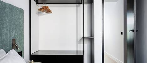 A modern hotel room with a bed, a small table with a glass, a hanging coat rack, and a door leading to another room.