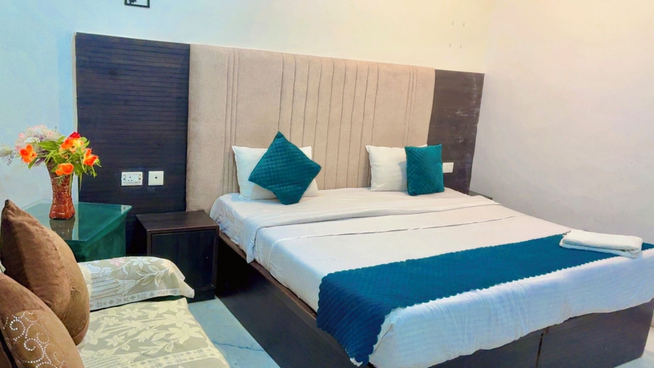 A hotel room with a bed, bedside tables, a nightstand, a vase with flowers, and a chair.
