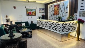 A reception area with a modern chandelier, a reception desk, green seating, and a decorative wall with framed pictures. - Hotel Taj Signature (Agra)
