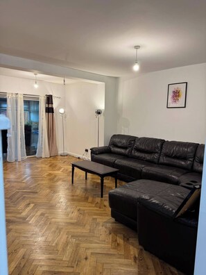 Un salon avec un canapé en cuir noir, un parquet en bois disposé en chevrons et une petite table basse noire. Il y a une fenêtre avec des rideaux légers et un lampadaire.