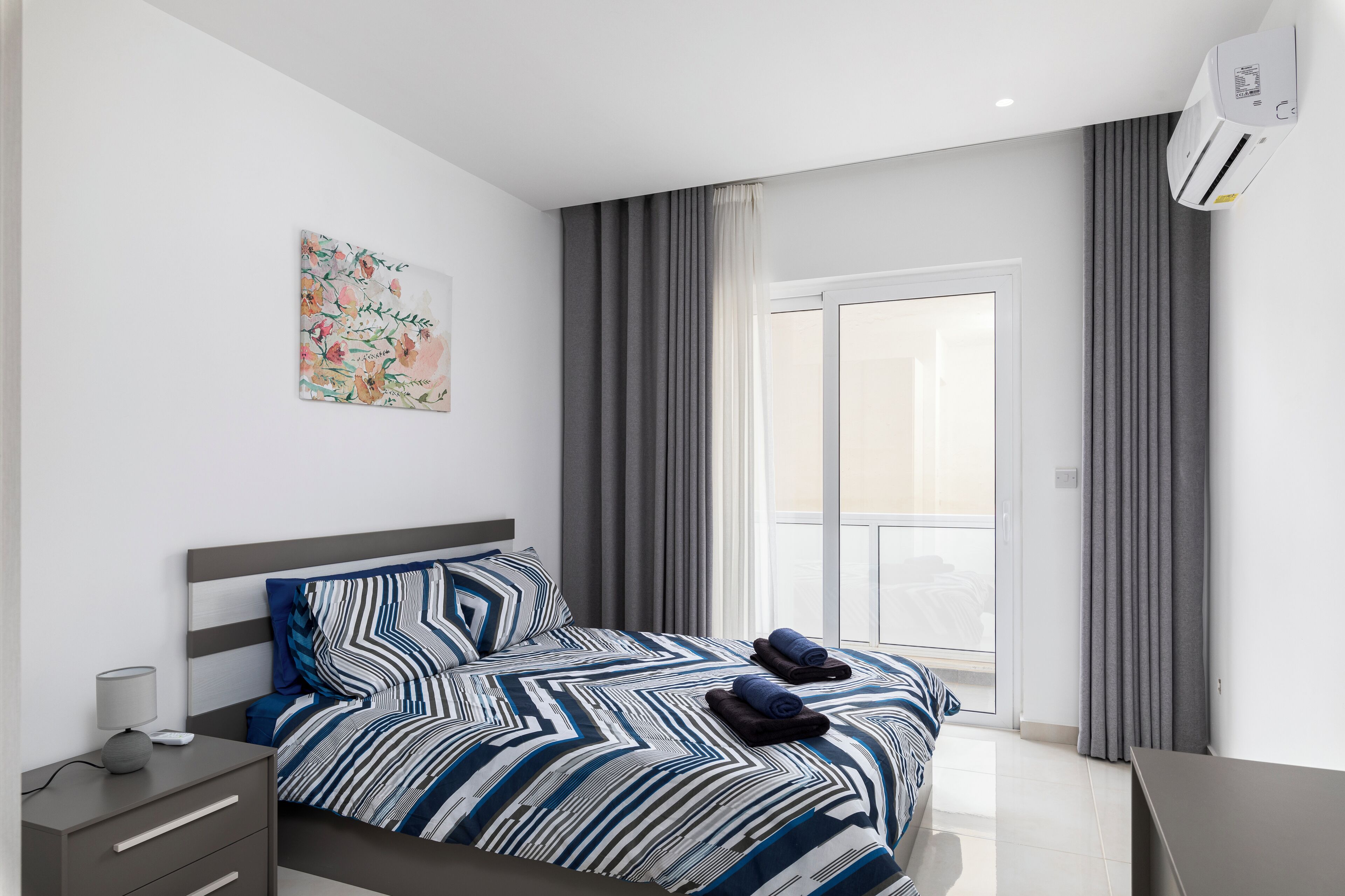 Une chambre d’hôtel moderne avec un grand lit, une table de chevet, une commode, une fenêtre avec des rideaux et un appareil de climatisation.