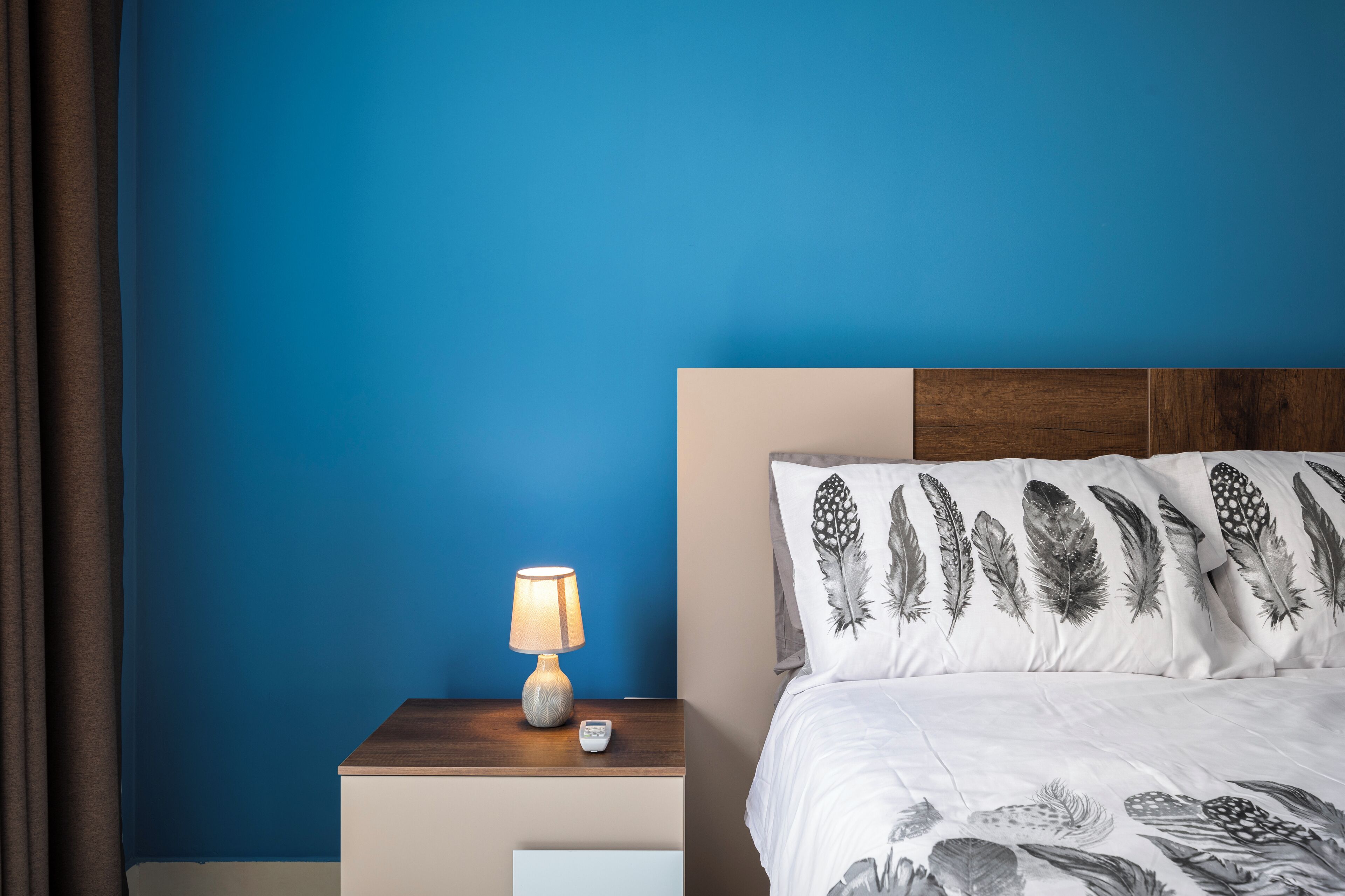 Ein Schlafzimmer mit blauer Wand, einem Nachttisch aus Holz mit Lampe und einem Bett mit Kissen mit Feder-Muster.