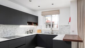 A modern kitchen with dark cabinets, a marble countertop, and a window with a view of buildings. - Modern Stylish 3 BR APT with Balcony (Swieqi)