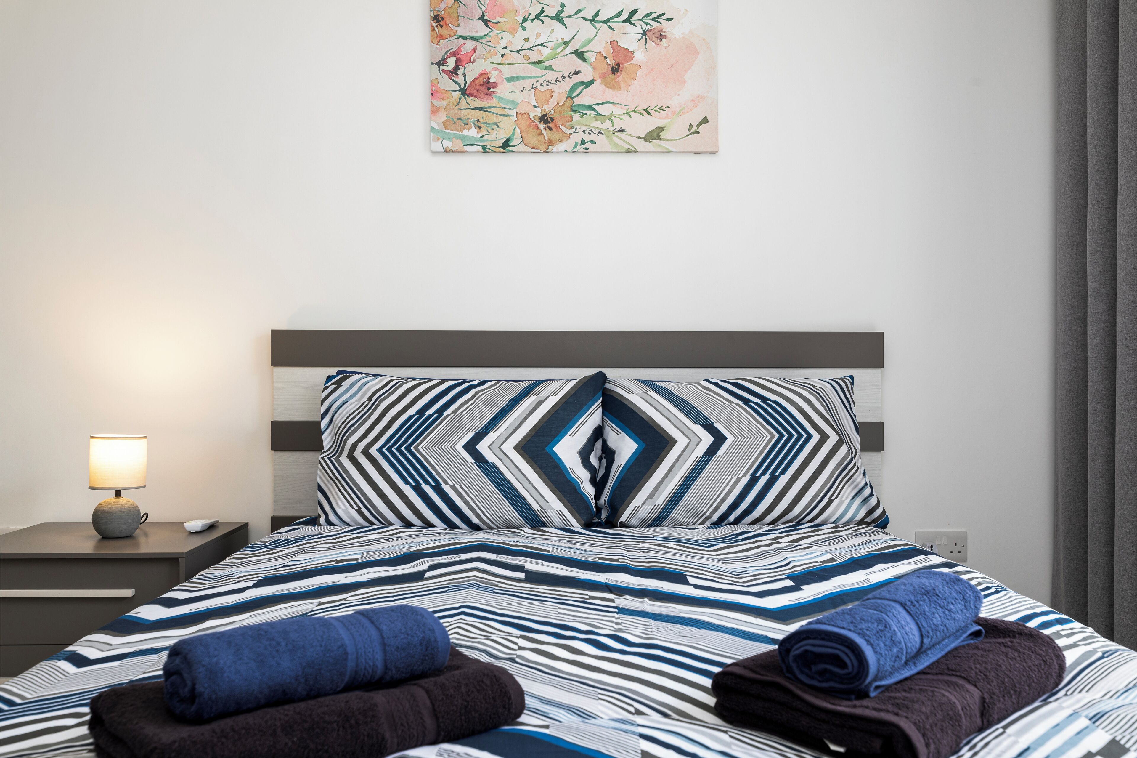 A neatly made bed with striped bedding and two pillows, a bedside lamp, and a floral artwork on the wall.