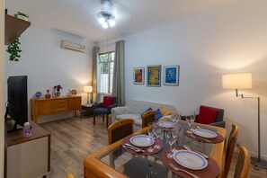 Un comedor con una mesa de vidrio dispuesta para cuatro personas, rodeada de sillas de madera. Se observa un sofá y un televisor al fondo.