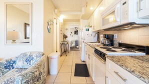 Cocina compacta con gabinetes blancos, estufa y microondas. Hay un cubo de basura negro y una pequeña mesa con una silla.
