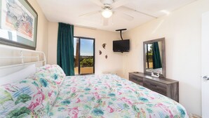 Un dormitorio con cama, ventilador de techo, televisor, cómoda, espejo y ventana con cortinas.