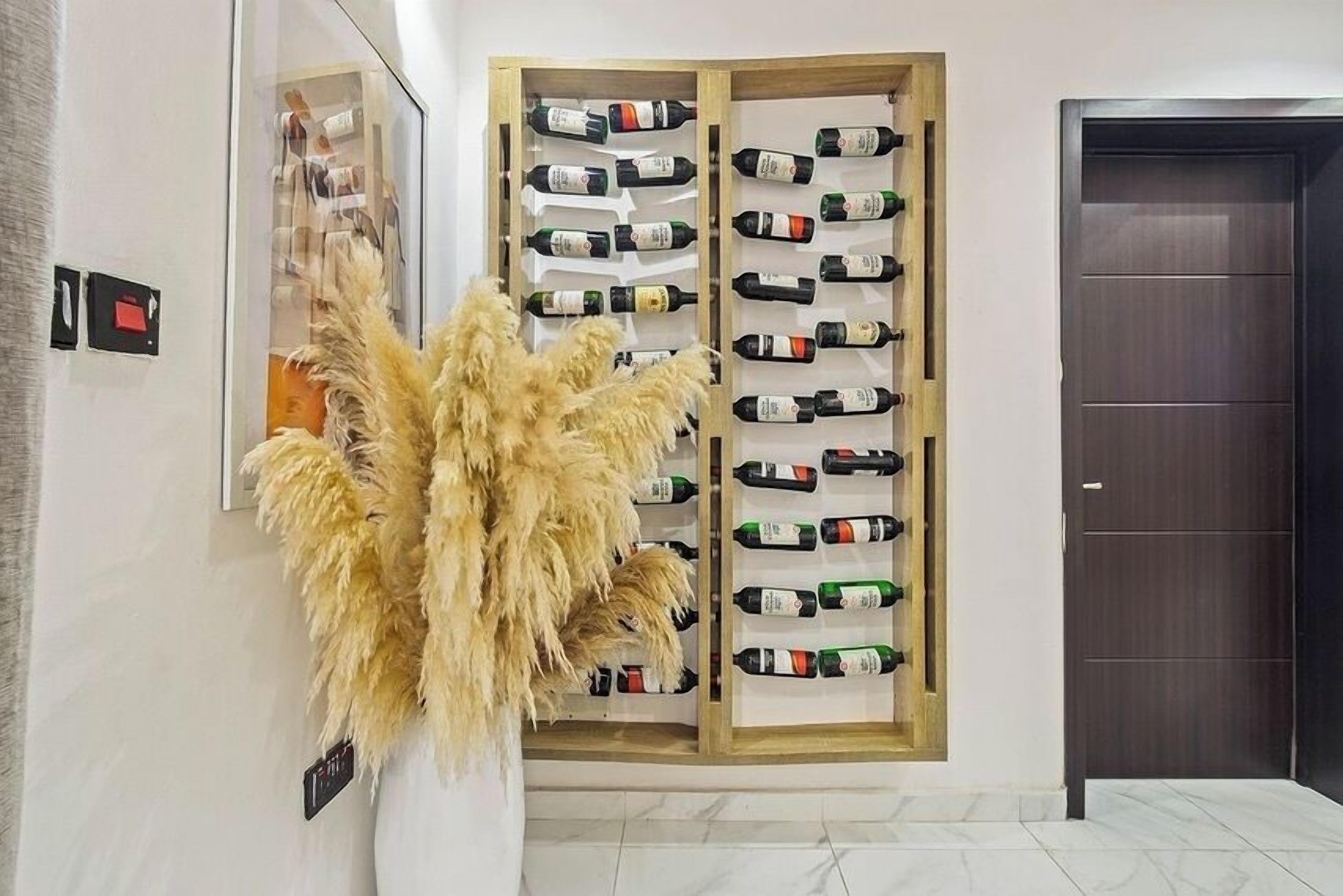 Una estantería para vino con varias botellas, una planta ornamental grande y una puerta de madera oscura en un pasillo.