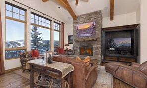A cozy living room with a stone fireplace, large windows with a snowy view, a television, and rustic furniture. - Big Sky Vacation Rentals: Big View Retreat (Big Sky)