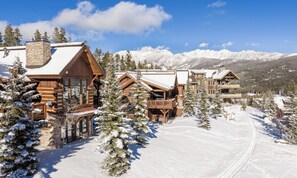 Ein schneebedecktes Chalet mit mehreren Holzhütten, einer Skipiste und einem Berg im Hintergrund.
