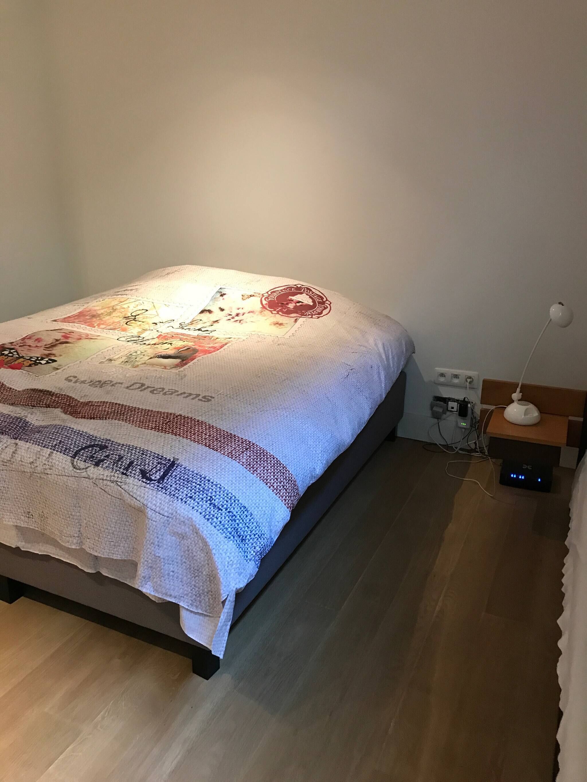 Un lit bien fait, recouvert d’une couverture à motifs, une table de chevet avec une lampe et un multiprise sur le sol.