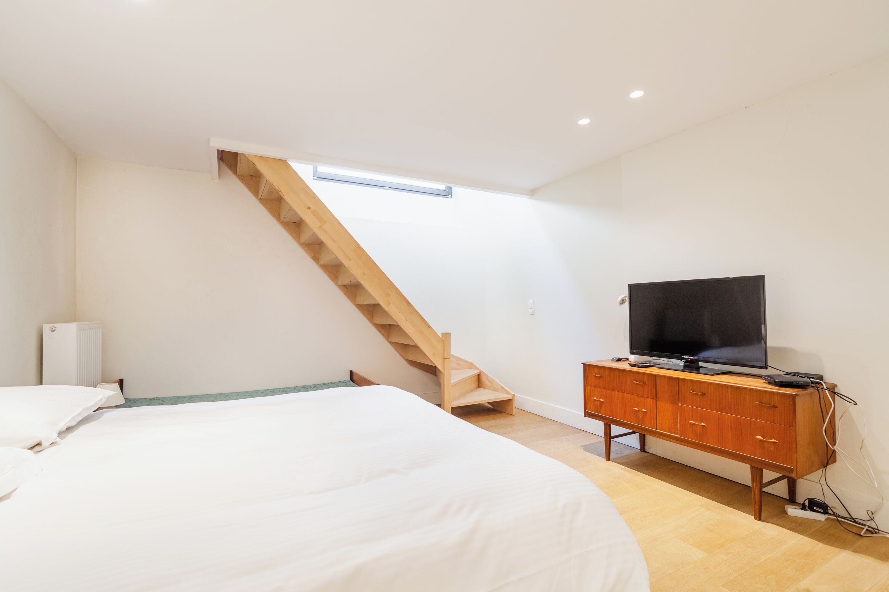A bedroom with a bed, a wooden dresser, a television, and a staircase.