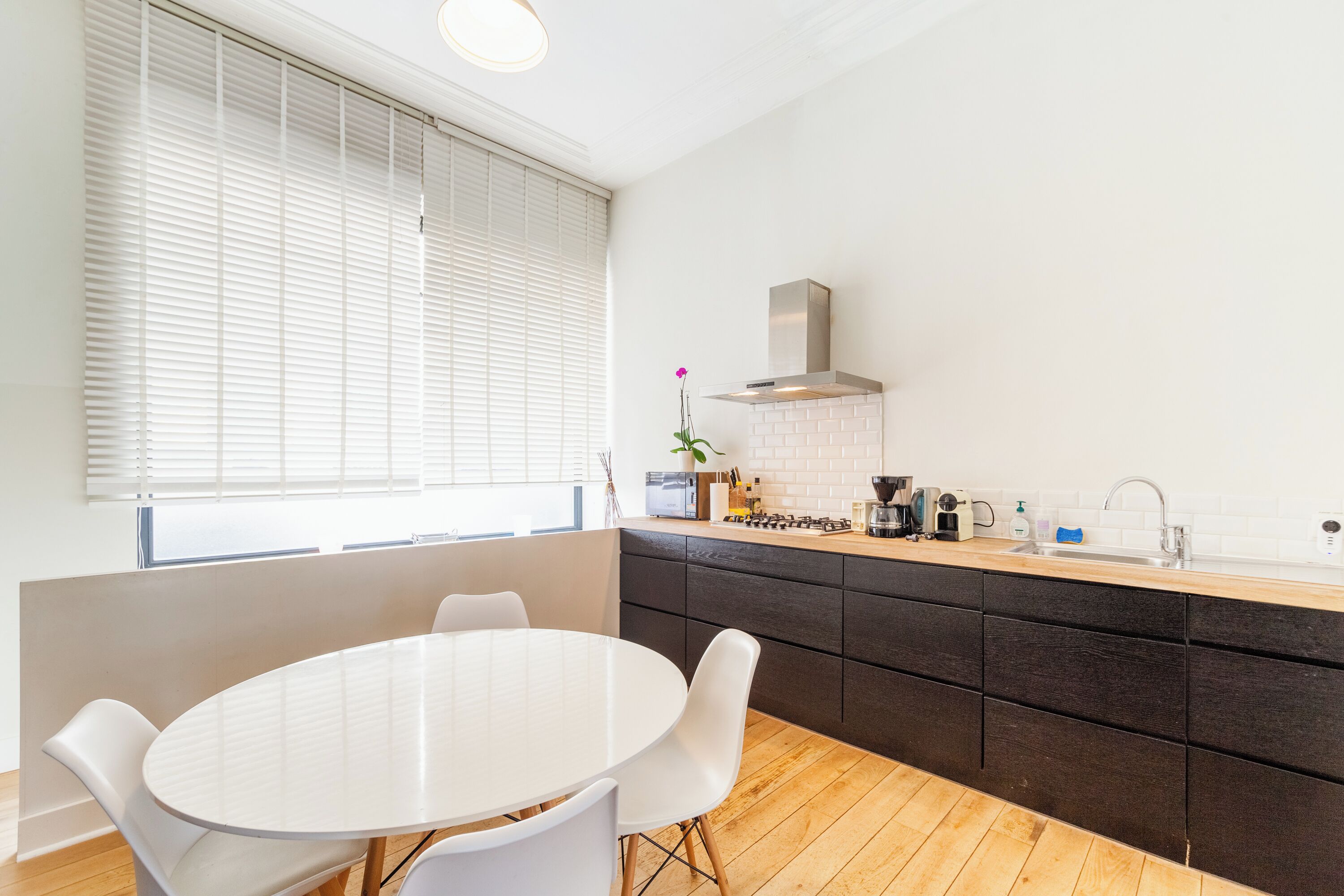 Une cuisine moderne avec une table et des chaises blanches, une cuisinière encastrée et une fenêtre avec des stores.