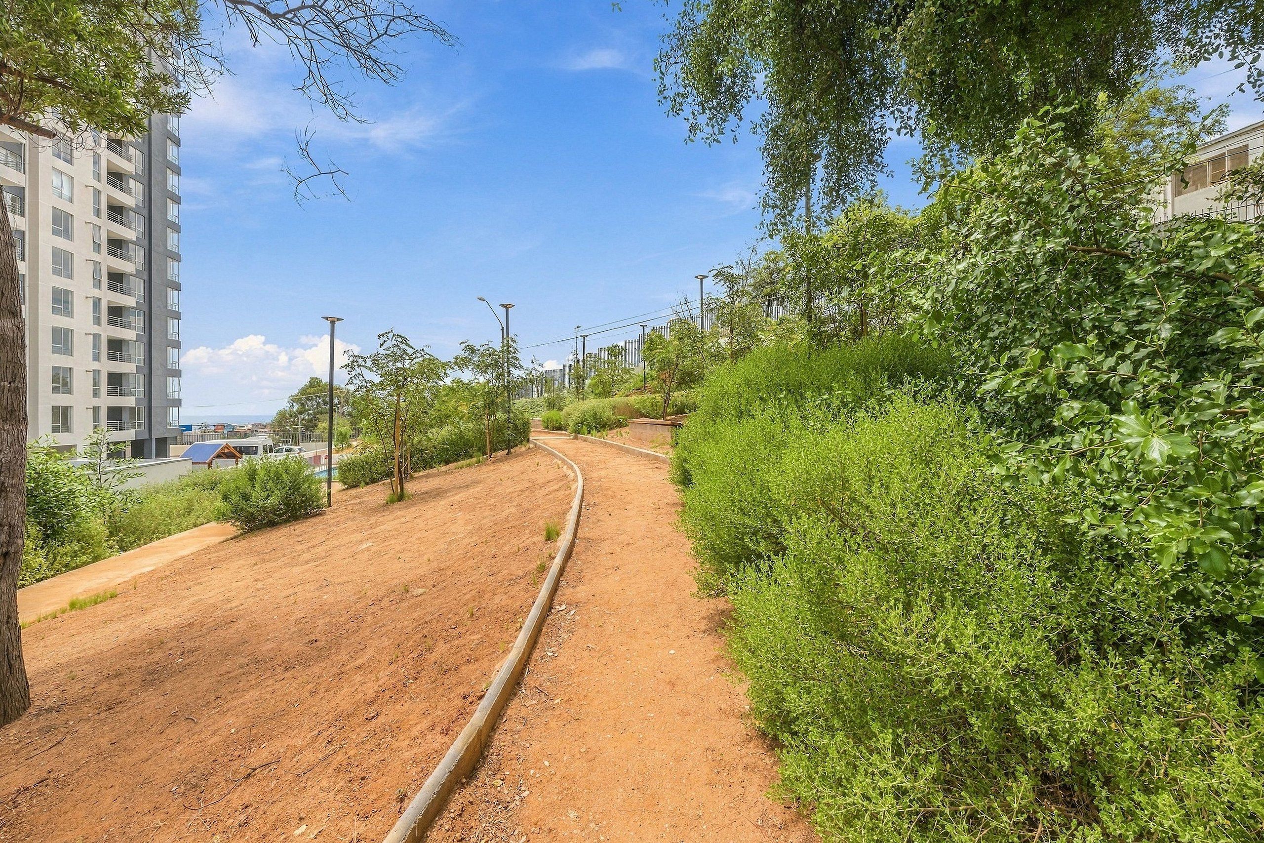 Camí de terra amb barana metàl·lica, envoltat de vegetació i un edifici al fons.