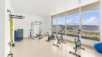 Un gimnasio con diversos equipos de ejercicio, una pelota de yoga y vistas a un paisaje urbano a través de amplias ventanas.