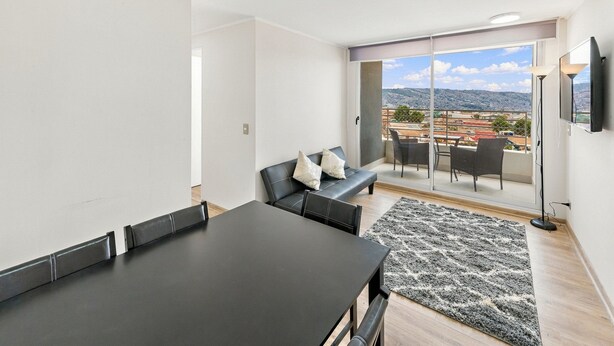 A modern living room with a dining area, a sofa with cushions, a large window with a city view, and a flat-screen TV mounted on the wall. - Oceana Suites Cerro Placeres (Valparaiso)