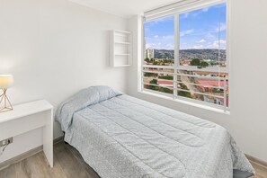 A bedroom with a bed, a desk, a lamp, and a window with a city view. - Oceana Suites Cerro Placeres (Valparaiso)