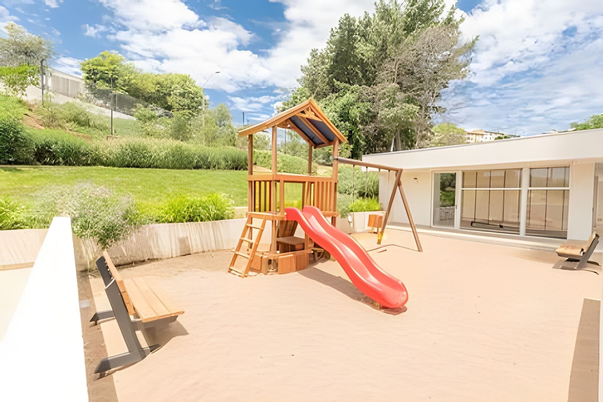 A playground with a wooden slide and climbing structure, surrounded by a landscaped garden and a building with large windows.