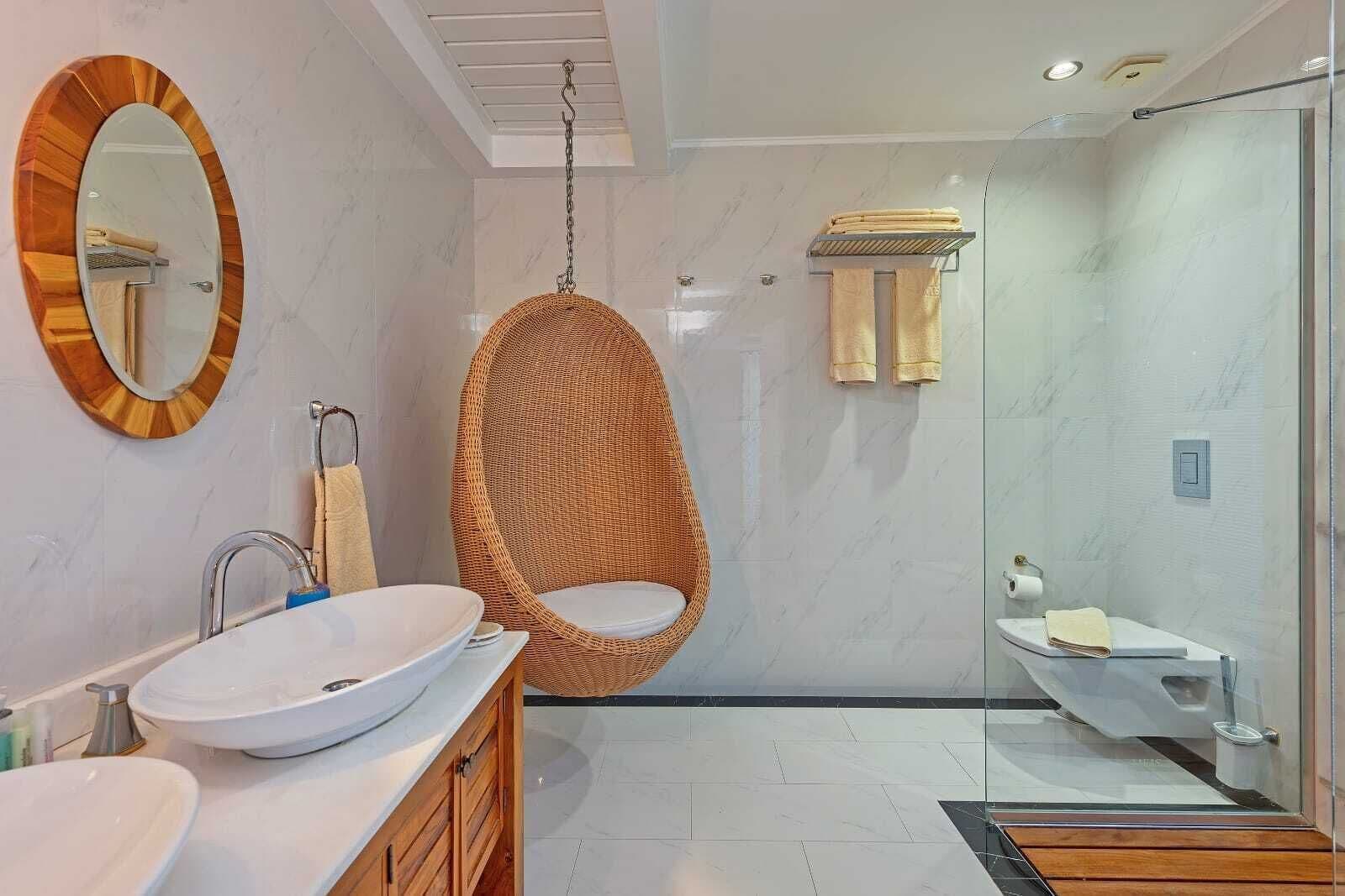 Une salle de bain moderne avec un grand miroir ovale, un fauteuil suspendu en osier, une douche en verre et un lavabo blanc doté d’un plan de travail en marbre.