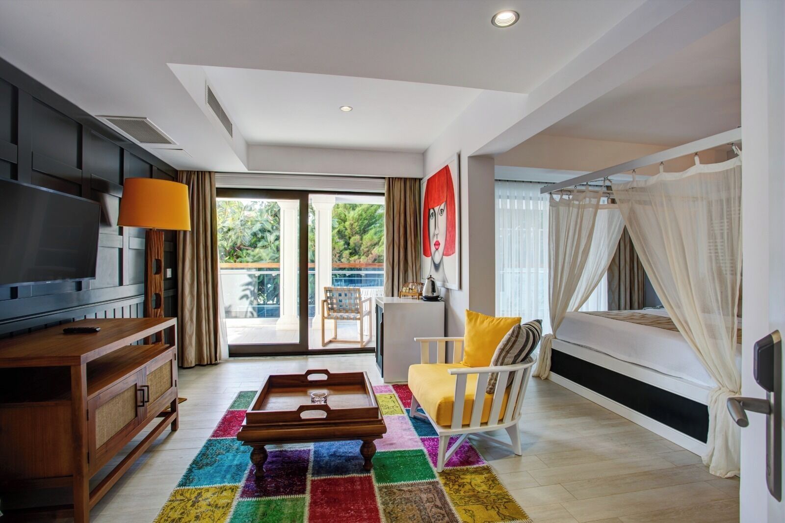 Una sala de estar moderna con televisor, muebles de madera, alfombra colorida, balcón con cortinas y un cuadro.