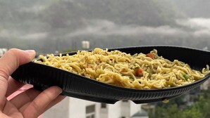 Una mano sosteniendo un recipiente negro para comida para llevar lleno de fideos y vegetales.