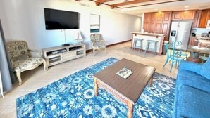 Una sala de estar con televisor, sofá y mesa de centro. Hay una zona de cocina con armarios de madera y una mesa de comedor.
