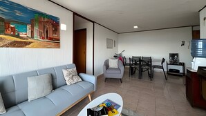A living room with a sofa, a small table with magazines, a painting on the wall, a dining area with chairs and a table, and a water dispenser. - Oceana Suites Valparaíso Yungay (Valparaíso)
