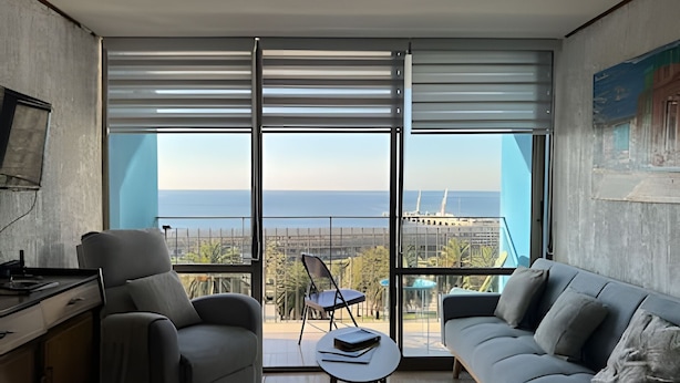 A modern living room with a sofa, chairs, a small table, and large windows offering a view of the sea and a distant building. - Oceana Suites Valparaíso Yungay (Valparaíso)