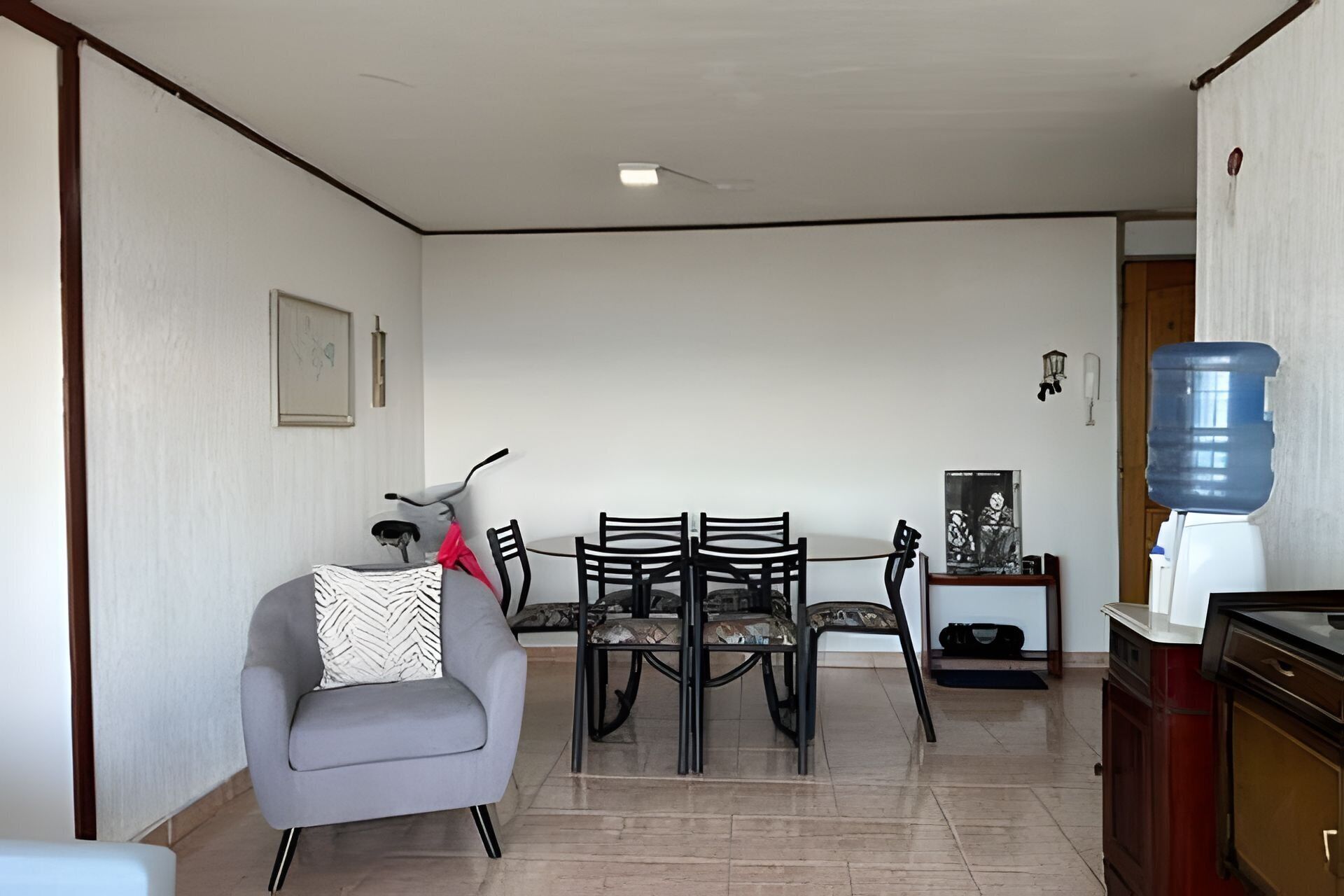 Una sala de estar con un sofá gris, una mesa de comedor con sillas, un dispensador de agua y un cuadro en la pared.