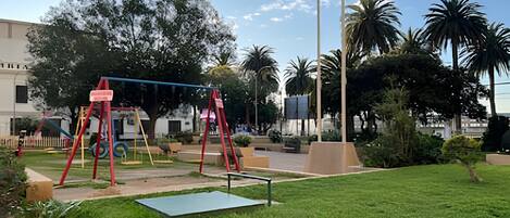 Un parque infantil con columpios y un tapete verde, rodeado de césped y palmeras.