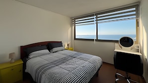 A bedroom with a bed, a bedside table, a desk with a chair, a window with blinds, and a view of the sea. - Oceana Suites Valparaíso Yungay (Valparaíso)