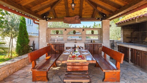 An outdoor kitchen with a stone countertop, wooden benches, and a pergola overhead.