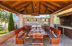 An outdoor kitchen with a stone countertop, wooden benches, and a pergola overhead. - Sea Mare Rustinella - A3 (Pula)