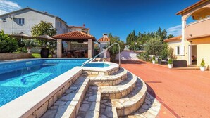 A swimming pool with stone steps, a covered seating area, and a patio with potted plants.