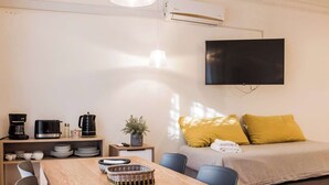 Ein modernes Zimmer mit einem Holztisch, schwarzen Stühlen, einer gelben Kissensofa sowie einem an der Wand befestigten Fernseher und einer Klimaanlage.