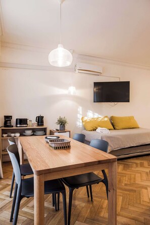 A modern room with a wooden dining table, black chairs, a sofa with yellow cushions, a mounted TV, and an air conditioning unit. - Metro Explorer near Hospital & Metro Baquedano (Providencia)