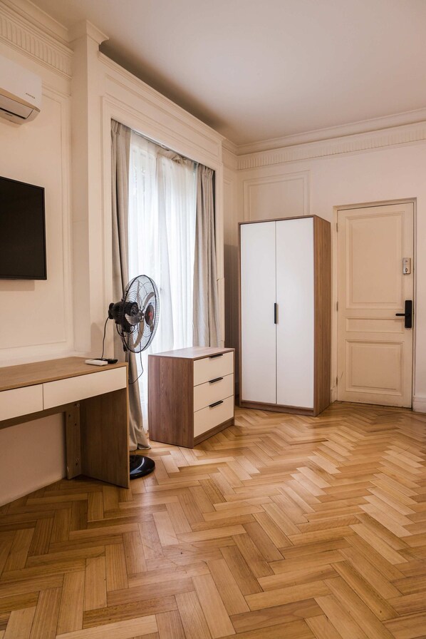 A room with a wooden herringbone floor, a desk with drawers, a standing fan, a wardrobe, and a door with a handle. - Elegant Studio | AC and Patio Perfect for Families (Providencia)